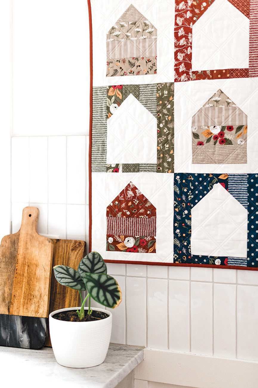 Quilted house wall hanging with cutting boards and plant.