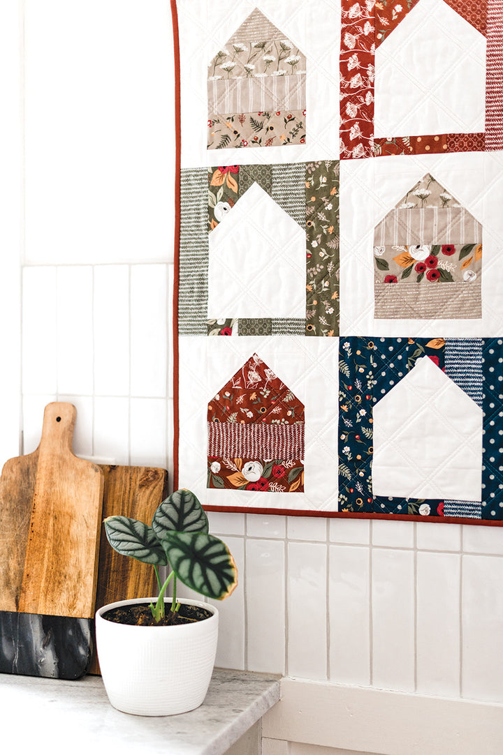 Quilted house wall hanging with cutting boards and plant.