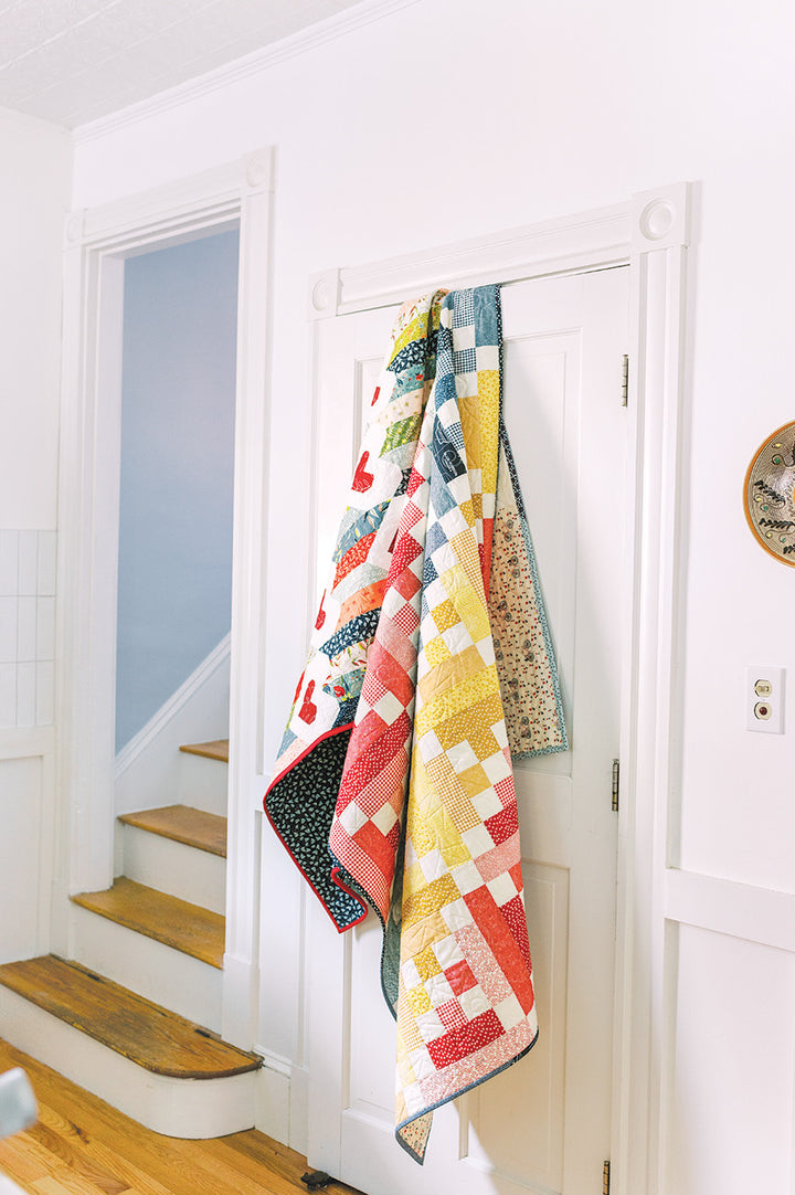 Quilts hanging on a white door, with stairs in the background.